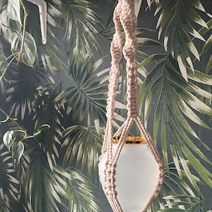 Puede incluir: Un macetero de macramé blanco con una maceta blanca con borde dorado que cuelga de una repisa blanca sobre un fondo de papel tapiz con estampado de hojas tropicales.