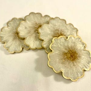 Puede incluir: Un juego de cinco posavasos con forma de flor. Cada posavasos tiene un borde dorado y un diseño de resina blanca y dorada. Los posavasos están colocados sobre una superficie blanca.