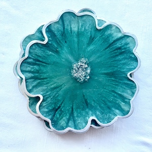 May include: A stack of teal resin coasters with a flower design. Each coaster has a silver-colored scalloped edge and a textured teal center with a silver glitter accent. The coasters are on a white surface.