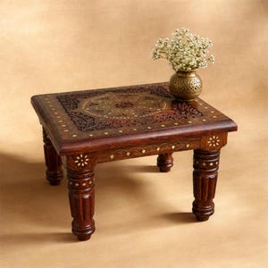 Puede incluir: Una mesa auxiliar rectangular de madera con acabado marrón oscuro e intrincados patrones de incrustaciones de latón en la parte superior y las patas. Un pequeño jarrón de latón redondo y perforado con flores blancas descansa sobre la mesa.