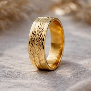 Könnte beinhalten: Ein goldfarbener Ring mit einem Weizenähren-Design. Der Ring hat ein breites Band mit geätzten Weizenähren. Der Ring liegt auf einer neutralfarbenen Oberfläche. Der Ring ist ein Schmuckstück.