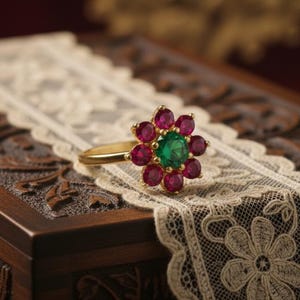 Puede incluir: Un anillo de oro con diseño floral, con una gema verde central rodeada de ocho gemas rojas. El anillo descansa sobre una superficie de madera decorativa con una superposición de encaje.
