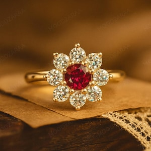 Peut inclure: Une bague en or avec un motif floral. La pierre centrale est rouge vif, entourée de huit pierres rondes et transparentes. La bague est montée sur un anneau en or, présentant un style classique et élégant.