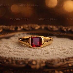 May include: A gold ring with a rectangular, deep red gemstone. The ring is set on a cream-colored, textured surface, and the background is blurred with warm tones. The ring's design is simple yet elegant, showcasing the vibrant gemstone.