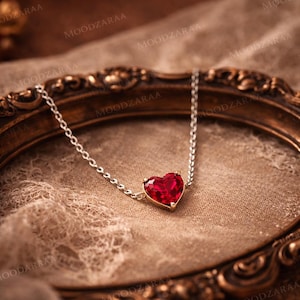Puede incluir: Collar de cadena plateada con un colgante en forma de corazón rojo rubí. El colgante está engastado en un bisel dorado. El collar se exhibe sobre una superficie texturizada, de color beige, con un marco decorativo dorado en el fondo.
