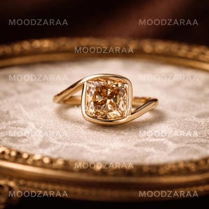 Op de afbeelding: Een gouden ring met een grote, vierkante, champagnekleurige edelsteen. De ring heeft een uniek, vloeiend ontwerp waarbij de band om de steen buigt. De ring ligt op een getextureerd, gebroken wit oppervlak.