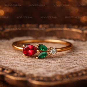 May include: A gold ring with a red, pear-shaped gemstone, two green leaf-shaped stones, and a small diamond. The ring is displayed on a textured surface. The text "MOODZARAA" is repeated.
