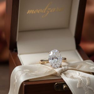 May include: An oval-cut diamond ring with a gold band sits in an open wooden jewellery box. The box is lined with cream fabric and tied with a cream ribbon. The word "moodzara" is printed in gold on the inside of the box.
