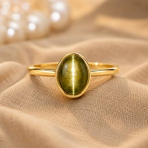 Op de afbeelding: Een gouden ring met een centrale ovaalvormige olijfgroene kattenoog-edelsteen. De gladde, gepolijste gouden band houdt de cabochon-steen vast in een eenvoudige zetting, wat zorgt voor een strakke en heldere uitstraling van dit sieraad.