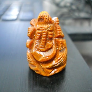 May include: A small, intricately carved wooden figurine in shades of brown and orange. The figure has a detailed design with visible textures and patterns, possibly depicting a religious or cultural symbol.