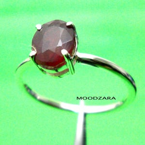 May include: A silver ring with a red gemstone set in a prong setting. The ring is on a green background.