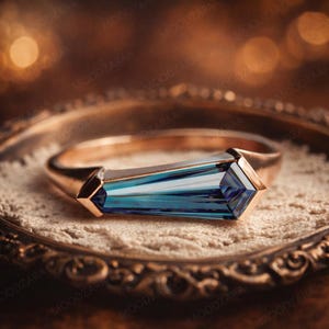 May include: A gold ring with a rectangular, blue gemstone. The gemstone has a faceted design, reflecting light. The ring is set on a decorative, gold-toned tray with a lace-like surface. The background is blurred with warm tones.