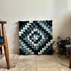 以下が含まれることがあります： 灰色と青の色合いの木製のブロックで作られた壁の芸術作品。ブロックは幾何学的なパターンで配置され、モダンで抽象的なデザインを生み出しています。芸術作品は白い壁にかけられており、手前に2つの椅子と植物があります。