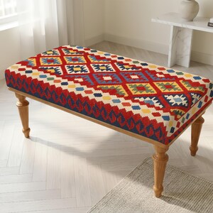 Op de afbeelding: Een rechthoekige voetenbank met een levendig, veelkleurig geometrisch patroon in rood, blauw, geel en groen. De gestoffeerde bovenkant rust op vier lichtgekleurde houten poten met een gedraaid, taps toelopend ontwerp voor een stabiele basis.