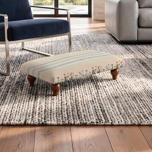 May include: A rectangular footstool with a cream-colored fabric top featuring light blue stripes and a pattern of small red and green floral motifs. The footstool is supported by four dark brown, rounded wooden legs with a turned design.