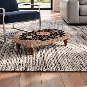 May include: A rectangular footstool featuring a low profile with four dark brown turned wooden feet. The top surface is upholstered in a fabric displaying a black, beige, and muted orange floral pattern reminiscent of a traditional rug design.