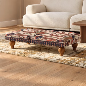 May include: A rectangular ottoman featuring a cream-colored fabric top with a repeating geometric pattern in shades of red, blue, and dark brown. The piece is supported by four short, rounded, dark wooden legs that elevate it from the floor.