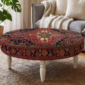 May include: A round ottoman featuring a detailed red, black, and cream-colored patterned textile top and sides. The piece is supported by four white, turned wooden legs that provide a stable base for the circular, upholstered furniture item.