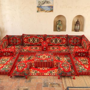 May include: A U-shaped floor seating set featuring a vibrant red base with intricate geometric patterns in green, yellow, and black. The set includes multiple floor cushions, backrest cushions, and smaller square pillows with decorative tassels.