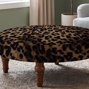 May include: A round ottoman featuring a brown and black leopard print fabric upholstery. The piece is supported by four turned wooden legs with a medium brown finish, providing a stable base for this circular furniture item.
