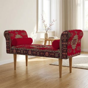 May include: A red upholstered bench featuring a detailed oriental rug pattern with intricate floral and geometric motifs. The bench has two matching cylindrical bolster pillows, curved armrests, and four light-coloured wooden legs.