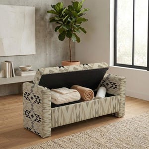 Könnte beinhalten: Eine rechteckige Aufbewahrungsbank mit geometrischem Muster in Beige-, Creme- und Schwarztönen. Der Klappdeckel ist geöffnet und gibt den Blick auf ein Innenfach frei, das eine gefaltete cremefarbene Strickdecke und eine gerollte beigefarbene Decke enthält.