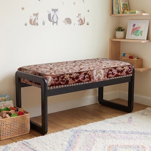 May include: A black bench with a rectangular seat upholstered in a deep red and cream fabric featuring an intricate floral damask pattern. The bench has a sturdy frame with rounded corners and four legs connected by horizontal support bars.