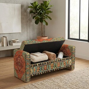 May include: A rectangular storage bench upholstered in a fabric featuring a multi-colored pattern of orange, teal, and black. The hinged lid is open, revealing an interior compartment containing a folded white knit blanket and a rolled tan blanket.