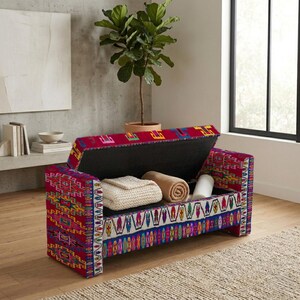 May include: A rectangular storage bench upholstered in a vibrant fabric featuring a multi-colored geometric pattern in shades of red, blue, yellow, and white. The hinged lid is open, revealing a hollow interior containing two folded blankets.