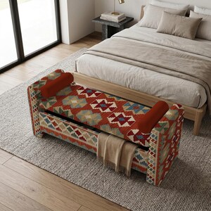 May include: A rectangular storage bench featuring a vibrant, multi-colored geometric pattern in shades of red, blue, and cream. The bench includes two solid rust-colored bolster pillows at each end and a beige throw blanket draped over the front.