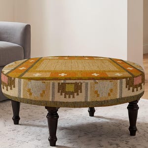 May include: A round ottoman featuring a woven fabric top and sides with a geometric pattern in shades of mustard yellow, orange, cream, and brown. The piece is supported by four dark brown turned wooden legs with a classic tapered design.
