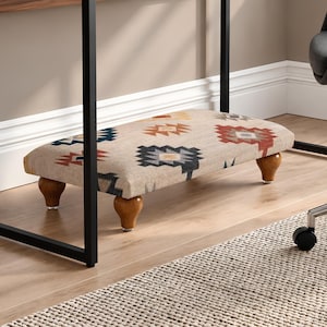 May include: A rectangular footstool with a beige fabric top featuring a geometric pattern in shades of dark blue, orange, and white. The footstool is supported by four short, rounded, brown wooden legs with a smooth, polished finish.