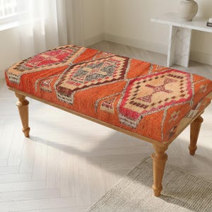 May include: A rectangular upholstered bench featuring a vibrant orange fabric with a repeating geometric pattern in shades of red, cream, and dark gray. The bench is supported by four turned wooden legs with a light brown finish and a smooth texture.