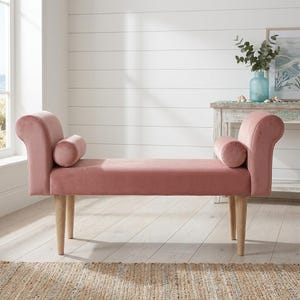 May include: A dusty rose velvet bench featuring rolled arms, two matching cylindrical bolster pillows, and four light-toned wooden legs. The bench has a smooth, upholstered seat and a structured frame with a soft, matte fabric finish.