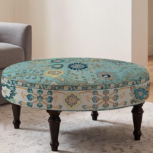 May include: A round ottoman featuring a teal fabric top with a multi-colored floral and geometric pattern. The side of the ottoman has a cream-colored band with a repeating blue and orange floral motif. It stands on four dark brown wooden legs.