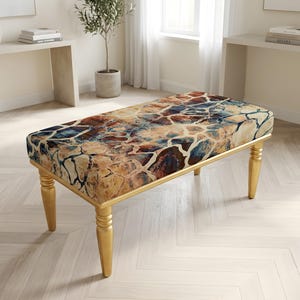 May include: A rectangular bench featuring a cushioned seat with a multi-colored abstract pattern of brown, tan, blue, and cream tones resembling cracked stone. The bench is supported by four tapered, gold-finished legs with a turned design.