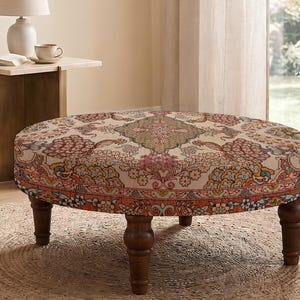 May include: A round ottoman featuring a beige fabric upholstery with a multi-colored intricate floral and paisley pattern in shades of red, orange, green, and blue. The piece is supported by four dark brown turned wooden legs in a classic style.