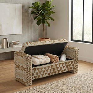 Puede incluir: Un banco de almacenamiento rectangular con un patrón geométrico en crema y bronceado, con una tapa abatible abierta. El compartimento interior contiene una manta de punto blanca doblada, una manta enrollada y un objeto cilíndrico blanco.