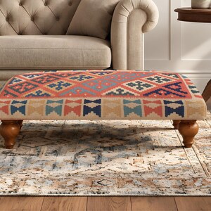 May include: A rectangular ottoman featuring a woven fabric top with a geometric pattern in shades of coral, navy blue, and beige. The piece is supported by four turned wooden legs with a warm brown finish, providing a stable base for the structure.