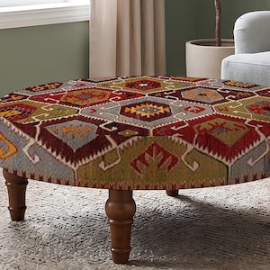 Puede incluir: Un otomán redondo con un patrón geométrico multicolor en tonos rojo, verde oliva, amarillo y gris. La tapicería tiene una textura tejida y la pieza se apoya sobre cuatro patas de madera marrón oscuro con un diseño torneado y ahusado.