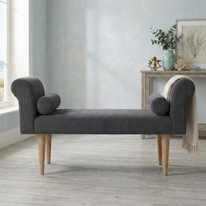 May include: A dark gray corduroy bench featuring a rolled arm design on both ends, two matching cylindrical bolster pillows, and four light-toned wooden legs. A cream-colored throw blanket is draped over the right side of the bench.
