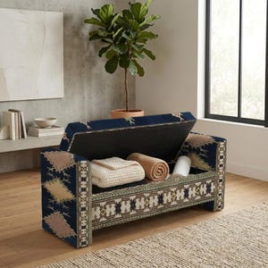 Puede incluir: Un banco de almacenamiento rectangular tapizado en tela azul oscuro con un patrón geométrico de diamantes multicolor. La tapa abatible está abierta, revelando un compartimento interior con una manta color crema doblada y una beige enrollada.