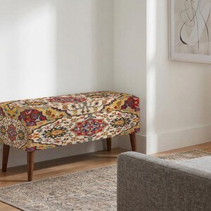 Könnte beinhalten: Eine rechteckige Aufbewahrungsbank mit einem lebendigen, mehrfarbigen Stoffmuster, das komplexe florale und geometrische Motive in Rot-, Gelb-, Blau- und Cremetönen aufweist und auf vier konisch geformten, dunkelbraunen Holzbeinen steht.