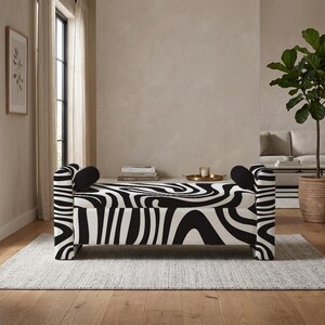 Puede incluir: Un banco de almacenamiento rectangular con un llamativo patrón ondulado en blanco y negro, similar al de una cebra, en el asiento y los laterales. El banco incluye dos pequeños cojines cilíndricos negros sólidos en cada extremo del asiento.