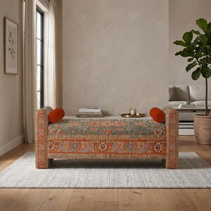 May include: A rectangular upholstered bench featuring a multi-colored floral and paisley pattern in shades of orange, teal, and cream. The bench includes two cylindrical orange bolster pillows placed at each end of the seat.