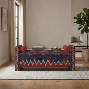 Puede incluir: Un banco rectangular tapizado con un vibrante patrón geométrico en tonos azul oscuro, rojo y blanco. El banco incluye dos cojines cilíndricos a juego en cada extremo, sostenidos por paneles laterales rectos y resistentes.