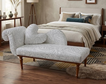 Puede incluir: Una chaise longue con tapizado de patrón de hojas botánicas en gris claro y blanco, respaldo curvo y un cojín decorativo en forma de corazón. El mueble se apoya sobre cuatro patas de madera marrón oscuro con diseño torneado.