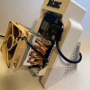 Könnte beinhalten: Ein weißes, 3D-gedrucktes Bitaxe-Gerät mit beigem Lüfter, Kupfer-Heatpipes und einer schwarzen Leiterplatte mit LCD-Bildschirm. Die Einheit ist auf einem strukturierten weißen Ständer mit Wabenmuster montiert und enthält ein schwarzes Stromkabel.