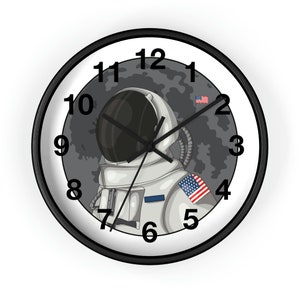 Puede incluir: Un reloj de pared blanco y negro con un astronauta en la luna. La esfera del reloj tiene números y manecillas negros. El astronauta lleva un traje espacial blanco con una bandera estadounidense en el hombro.