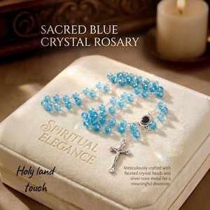 May include: A rosary featuring faceted blue crystal beads and silver-tone metal links, including a crucifix and a center piece with a black oval inlay. The rosary rests on a cream-colored box with the text Sacred Blue Crystal Rosary and Spiritual Elegance.